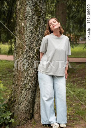 Woman in a gray t-shirt and light jeans leans against a tree with closed eyes, enjoying the moment in a green park Woman in a gray t-shirt and light jeans leans against a tree with closed eyes, enjoying the moment in a green park 131406600