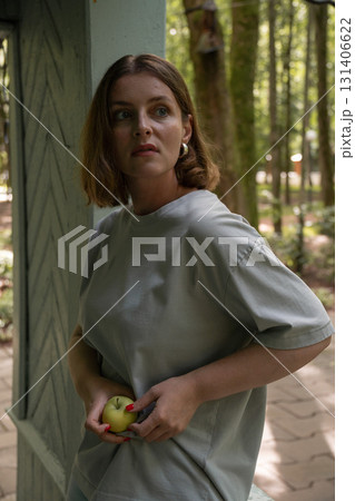 Woman in a gray t-shirt holds a green apple while looking thoughtfully to the side in a park pavilion in Peredelkino Woman in a gray t-shirt holds a green apple while looking thoughtfully to the side in a park pavilion in Peredelkino 131406622