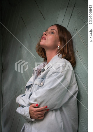 A thoughtful young woman in a light denim jacket leans against a rustic wooden wall, gazing upwards in a pensive pose A thoughtful young woman in a light denim jacket leans against a rustic wooden wall, gazing upwards in a pensive pose 131406642