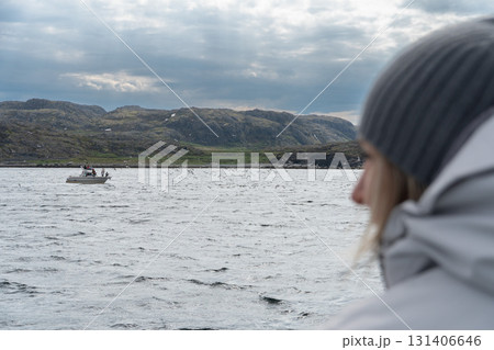 A person in a white jacket and gray knitted hat looks at small boats on the Barents Sea under a cloudy sky A person in a white jacket and gray knitted hat looks at small boats on the Barents Sea under a cloudy sky 131406646