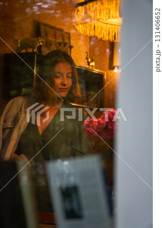 A woman in a green dress is seen through a window in a cozy vintage room with warm lighting and a bouquet of flowers. Peredelkino A woman in a green dress is seen through a window in a cozy vintage room with warm lighting and a bouquet of flowers. Peredelkino 131406652