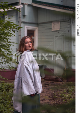 A close-up portrait of a woman in a green dress with a denim jacket draped over her shoulders standing near a pastel-colored wooden house 131406654