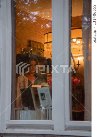 A young woman is seen through a window, reading a book in a cozy vintage-style room with warm lighting and retro decor A young woman is seen through a window, reading a book in a cozy vintage-style room with warm lighting and retro decor 131406655