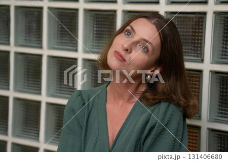 Close-up portrait of a woman in a green dress against a glass block wall, looking upward with a thoughtful expression Close-up portrait of a woman in a green dress against a glass block wall, looking upward with a thoughtful expression 131406680