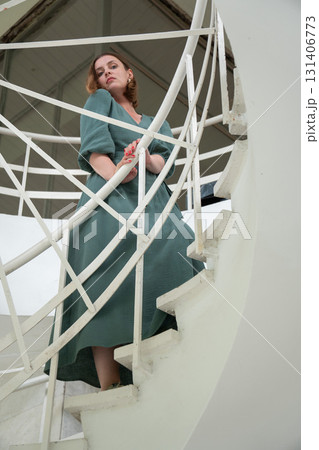 Woman in a green dress leans on a white spiral staircase, captured from a low angle with graphic architectural lines 131406773
