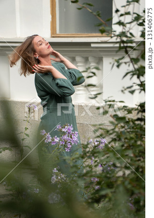 A woman in a green dress poses gracefully among purple flowers against a white building wall, her hair moving in the air A woman in a green dress poses gracefully among purple flowers against a white building wall, her hair moving in the air 131406775