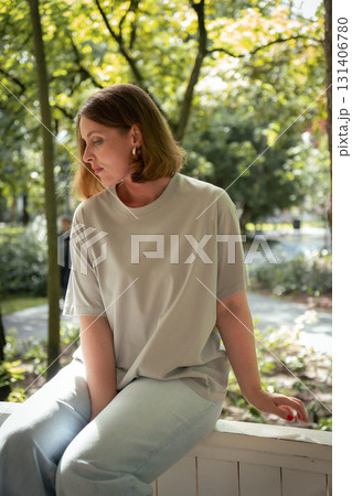 Woman in a beige T-shirt and light jeans sits thoughtfully on a wooden railing in a sunlit park 131406780