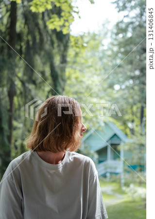 Woman in casual clothes sitting outdoors and looking to the side, with a green wooden house in the blurred background and forest surroundings Woman in casual clothes sitting outdoors and looking to the side, with a green wooden house in the blurred background and forest surroundings 131406845