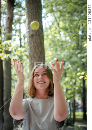 Smiling woman with red nails tosses a green apple in the air while standing in a sunlit forest in Peredelkino 131406846