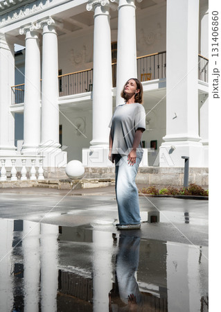 Woman in casual clothes stands near a neoclassical building, reflected in a large puddle on the pavement after rain Woman in casual clothes stands near a neoclassical building, reflected in a large puddle on the pavement after rain 131406860