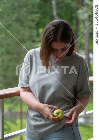 Young woman in light gray t-shirt holding a green apple and looking down thoughtfully while standing on a wooden balcony surrounded by lush greenery Young woman in light gray t-shirt holding a green apple and looking down thoughtfully while standing on a wooden balcony surrounded by lush greenery 131406928