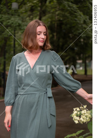 A woman in a green wrap dress stands on a park path, gently touching blooming white hydrangeas 131406955