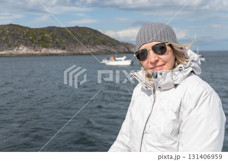 Woman in white jacket and gray knit hat smiles on a boat, rocky coastline and other vessels in the background, clear sky and cold wind 131406959