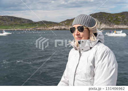 Woman in white jacket and gray knit hat smiles on a boat, rocky coastline and other vessels in the background, clear sky and cold wind Woman in white jacket and gray knit hat smiles on a boat, rocky coastline and other vessels in the background, clear sky and cold wind 131406962
