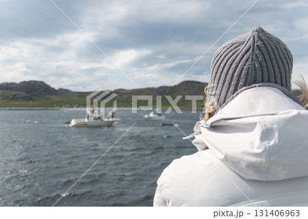 A person in a white jacket and gray knitted hat looks at small boats on the Barents Sea under a cloudy sky A person in a white jacket and gray knitted hat looks at small boats on the Barents Sea under a cloudy sky 131406963