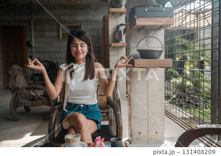 A young woman meditates in a stylish, industrial-themed home, surrounded by natural light and cozy decor. 131408209