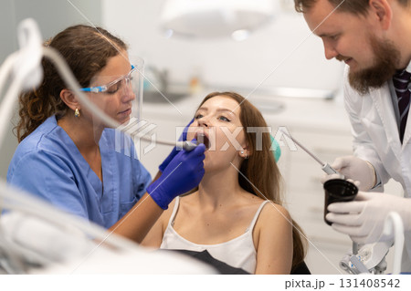 Girl is sitting in armchair during dental treatment at dentist appointment. Female doctor performs procedure of restoration and improvement of teeth, uses technological device Girl is sitting in armchair during dental treatment at dentist appointment. Female doctor performs procedure of restoration and improvement of teeth, uses technological device 131408542