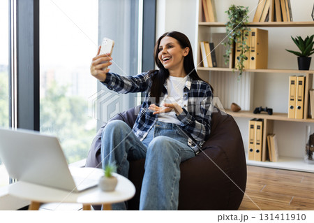 Young woman in her 20s having an online video chat using smartphone, sitting in a cozy room on a bean bag chair next to window. She seems cheerful and enjoying communication. 131411910