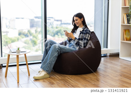 Young adult woman seated on bean bag in modern lounge environment using smartphone. Scene shows casual lifestyle and leisure time indoors. Well-lit surroundings with natural light and wooden flooring. Young adult woman seated on bean bag in modern lounge environment using smartphone. Scene shows casual lifestyle and leisure time indoors. Well-lit surroundings with natural light and wooden flooring. 131411921