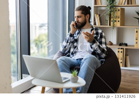 Man in his 30s speaking on phone while working remotely, using laptop and sitting at home 131411926