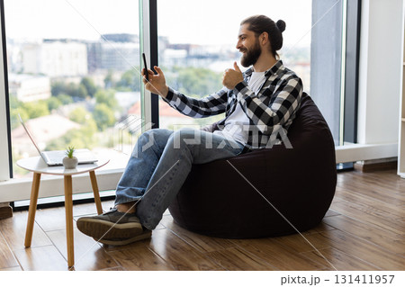 Young adult male sitting on beanbag chair in office, chatting through smartphone during workday. Window view in bright modern space portrays casual, multitasking approach to remote communication. 131411957