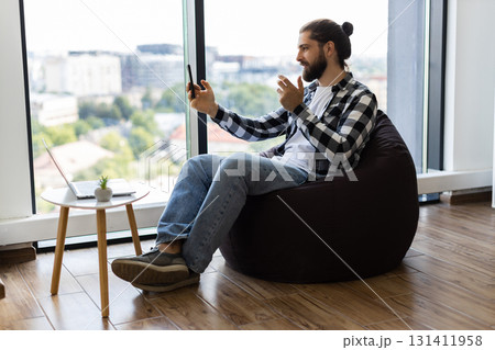 Young adult male sitting on beanbag chair in office, chatting through smartphone during workday. Window view in bright modern space portrays casual, multitasking approach to remote communication. Young adult male sitting on beanbag chair in office, chatting through smartphone during workday. Window view in bright modern space portrays casual, multitasking approach to remote communication. 131411958