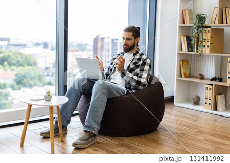 Middle-aged man with beard and laptop engaged in video call, sitting in cozy living room 131411992