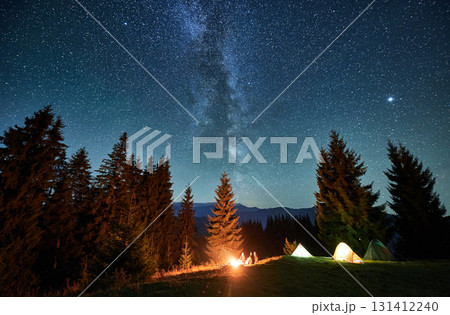 Panoramic view of grassy hill with illuminated camp tents and coniferous trees under starry sky. Night camping in mountains under beautiful sky with stars. Concept of hiking, nighttime and galaxy. Panoramic view of grassy hill with illuminated camp tents and coniferous trees under starry sky. Night camping in mountains under beautiful sky with stars. Concept of hiking, nighttime and galaxy. 131412240