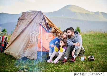 Family of three sits together in front of orange tent in grassy mountain campsite. Child uses binoculars while parents share tender moment. Peaceful and intimate outdoor experience. Family of three sits together in front of orange tent in grassy mountain campsite. Child uses binoculars while parents share tender moment. Peaceful and intimate outdoor experience. 131412245