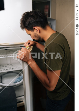 Vertical shot of focused young man cleaning refrigerator door seal with yellow sponge, focusing on detailed sanitation and preserving appliance functionality during routine kitchen maintenance. 131417916