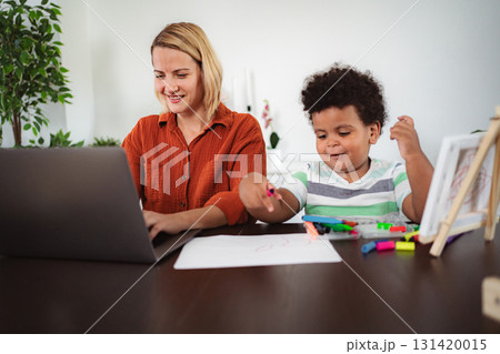 Mother working on laptop with child drawing at home Mother working on laptop with child drawing at home 131420015