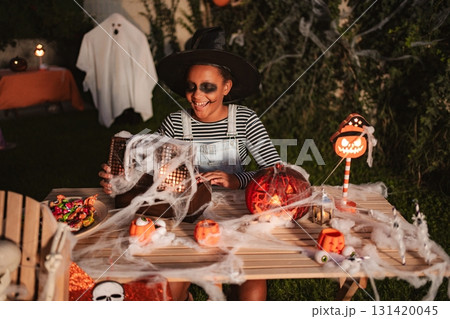 Girl wearing witch costume looking at Halloween decorations on a table in the garden Girl wearing witch costume looking at Halloween decorations on a table in the garden 131420045