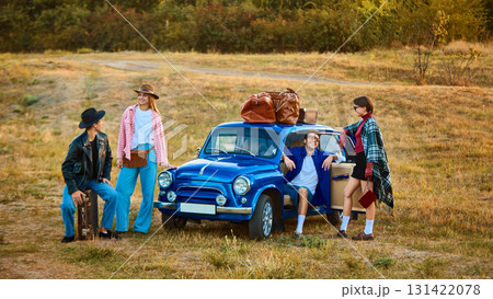 Group of friends in retro style outfits standing by blue car with luggage on roof Group of friends in retro style outfits standing by blue car with luggage on roof 131422078