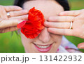 A woman playfully hides her eye with a vibrant red poppy flower, smiling gently 131422932