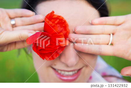A woman playfully hides her eye with a vibrant red poppy flower, smiling gently A woman playfully hides her eye with a vibrant red poppy flower, smiling gently 131422932