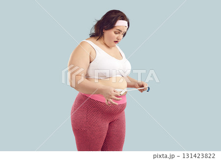 Overweight woman with fat belly looks at measuring tape with surprised face expression Overweight woman with fat belly looks at measuring tape with surprised face expression 131423822