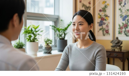 Asian woman in gray ribbed sweater having warm conversation with friend in cozy home interior with botanical artwork and green plants by sunny window 131424193