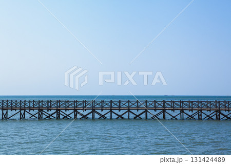 Long wooden bridge stretches across calm turquoise water. Traditional Indonesian design, serene seascape, clear blue sky create peaceful tropical atmosphere, ideal for travel and tourism promotions. Long wooden bridge stretches across calm turquoise water. Traditional Indonesian design, serene seascape, clear blue sky create peaceful tropical atmosphere, ideal for travel and tourism promotions. 131424489