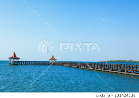 People walk by long wooden pier with traditional gazebos stretches over calm turquoise sea under clear blue sky. Perfect tropical scenery for travel, resort, summer vacation, coastal tourism themes. People walk by long wooden pier with traditional gazebos stretches over calm turquoise sea under clear blue sky. Perfect tropical scenery for travel, resort, summer vacation, coastal tourism themes. 131424539