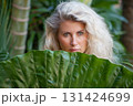 Blonde woman hiding behind a giant tropical leaf in a lush jungle in Bali. Natural portrait that evokes travel, wellness, connection with nature. Ideal for spa, retreats, summer travel lifestyle. 131424699
