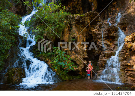 Mother with child standing under scenic Goa Tetes waterfall in East Java, Indonesia. Lush jungle surroundings, dramatic cliffs, and cascading streams create perfect backdrop for adventurous family Mother with child standing under scenic Goa Tetes waterfall in East Java, Indonesia. Lush jungle surroundings, dramatic cliffs, and cascading streams create perfect backdrop for adventurous family 131424704