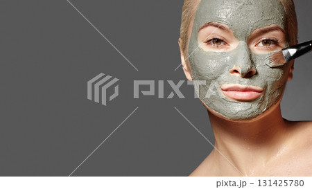Close-up portrait of beautiful woman applying gray clay facial mask for skincare and beauty treatment. Spa girl with natural fresh skin, holding cosmetic product brush. Organic cosmetics, anti-aging Close-up portrait of beautiful woman applying gray clay facial mask for skincare and beauty treatment. Spa girl with natural fresh skin, holding cosmetic product brush. Organic cosmetics, anti-aging 131425780