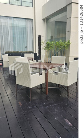A tranquil outdoor dining area features several modern tables and wicker chairs arranged on a dark wooden deck, surrounded by lush green plants and a contemporary building. 131426684