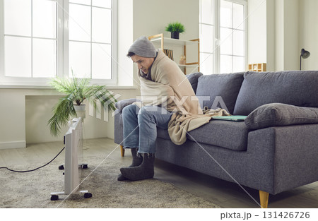 Man sitting on sofa at home wrapped in blanket and trying to warm up on electric heater. Man sitting on sofa at home wrapped in blanket and trying to warm up on electric heater. 131426726