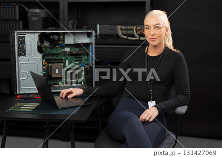 Female IT technician in her 30s with laptop beside open server, performing hardware maintenance and diagnostics Female IT technician in her 30s with laptop beside open server, performing hardware maintenance and diagnostics 131427069