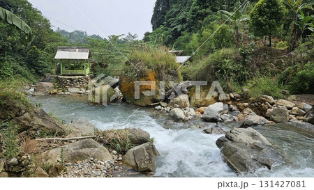 This lush tropical riverscape showcases cascading clear water rushing over boulders and pebbles in a rocky streambed, framed by dense green vegetation and tall trees, with a small, rustic wooden shelt This lush tropical riverscape showcases cascading clear water rushing over boulders and pebbles in a rocky streambed, framed by dense green vegetation and tall trees, with a small, rustic wooden shelt 131427081