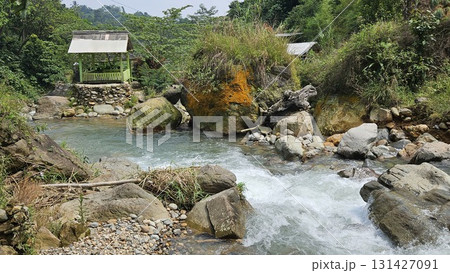 This lush tropical riverscape showcases cascading clear water rushing over boulders and pebbles in a rocky streambed, framed by dense green vegetation and tall trees, with a small, rustic wooden shelt 131427091