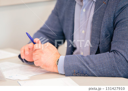 Hands of an unrecognizable male businessman at the table. He is wearing a blue suit and holding a pen in his hands. Hands of an unrecognizable male businessman at the table. He is wearing a blue suit and holding a pen in his hands. 131427910