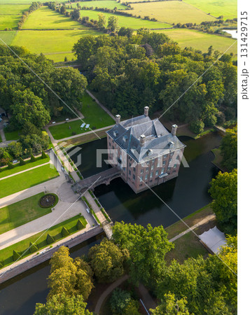 Aerial view of historic moated castle with gardens, bridge, and adjacent long building, surrounded by trees, water features, and a nearby village under warm sunlight. 131429715