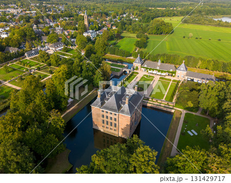 Aerial view of historic moated castle with gardens, bridge, and adjacent long building, surrounded by trees, water features, and a nearby village under warm sunlight. Aerial view of historic moated castle with gardens, bridge, and adjacent long building, surrounded by trees, water features, and a nearby village under warm sunlight. 131429717